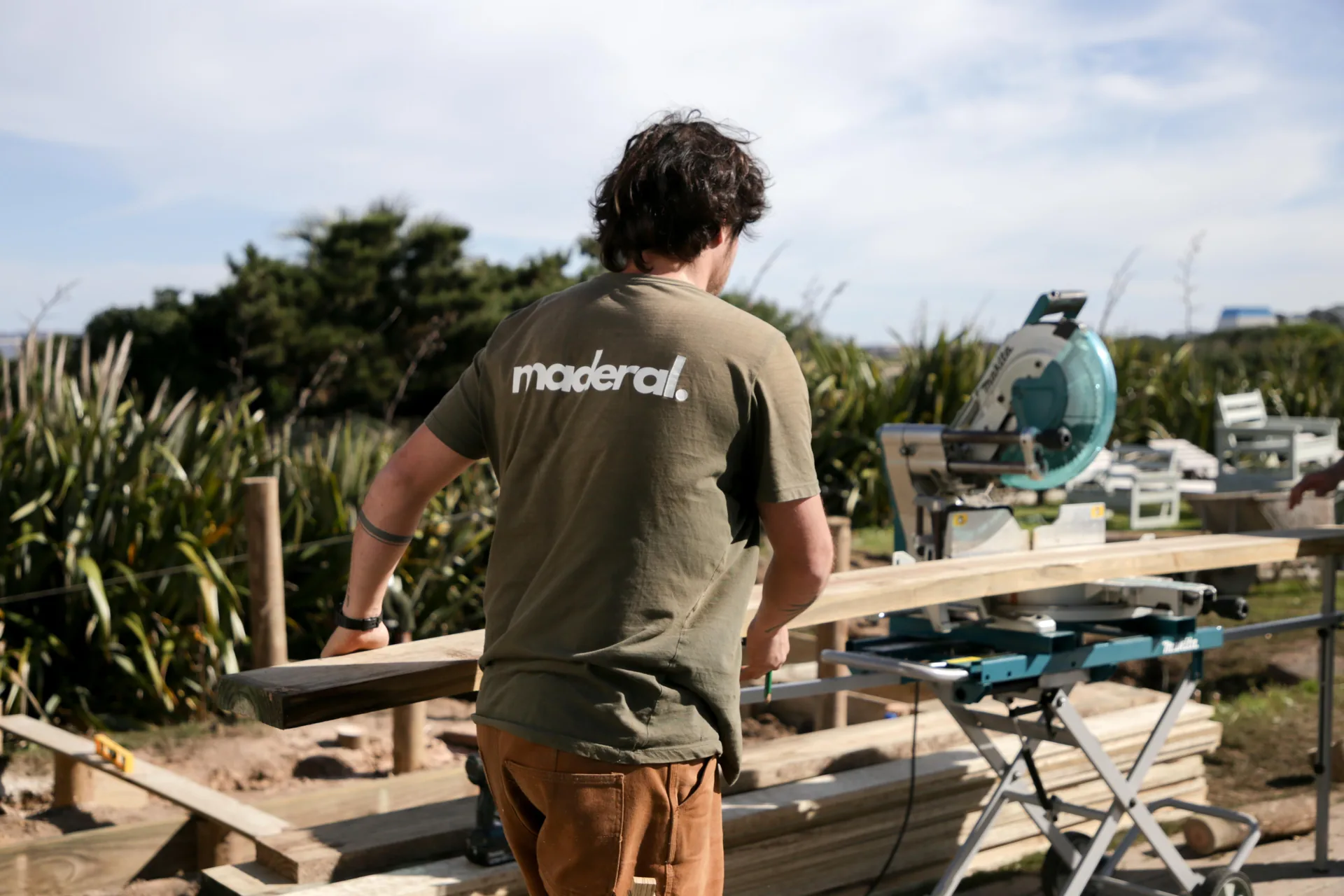 Carpintero con remera de Maderal junto a una sierra y tablas de madera al aire libre